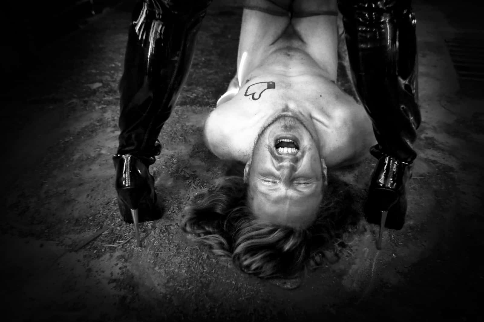 Mathias naked on the floor between the tall boots of a dominant woman.