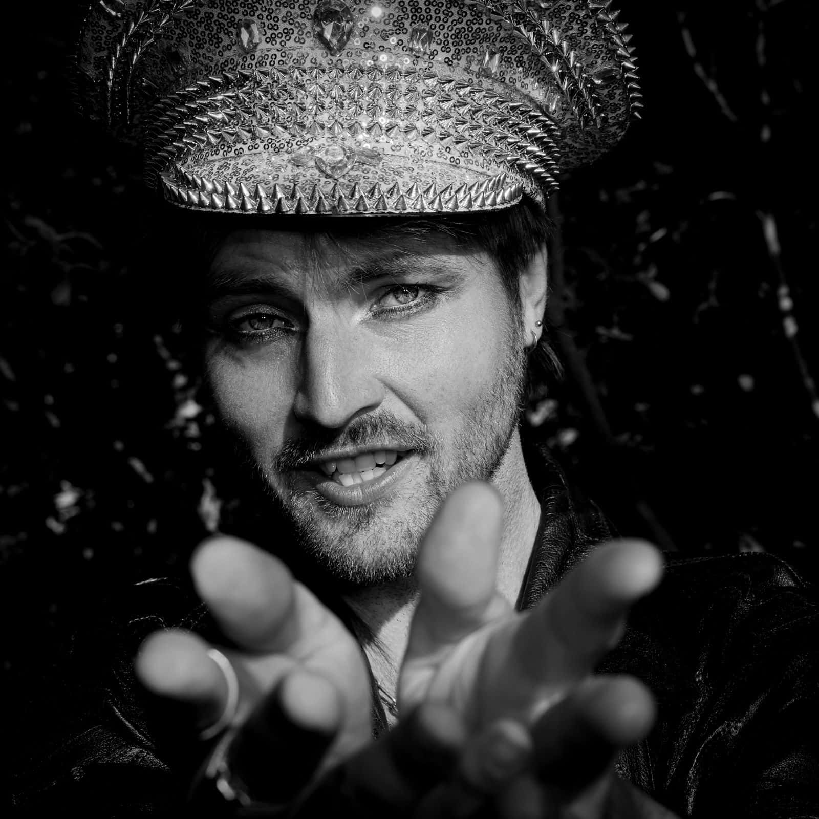 Johan looks directly into the camera, wearing an ornate cap and dark clothing, with his hand reaching toward the viewer in a playful, intimate portrait.
