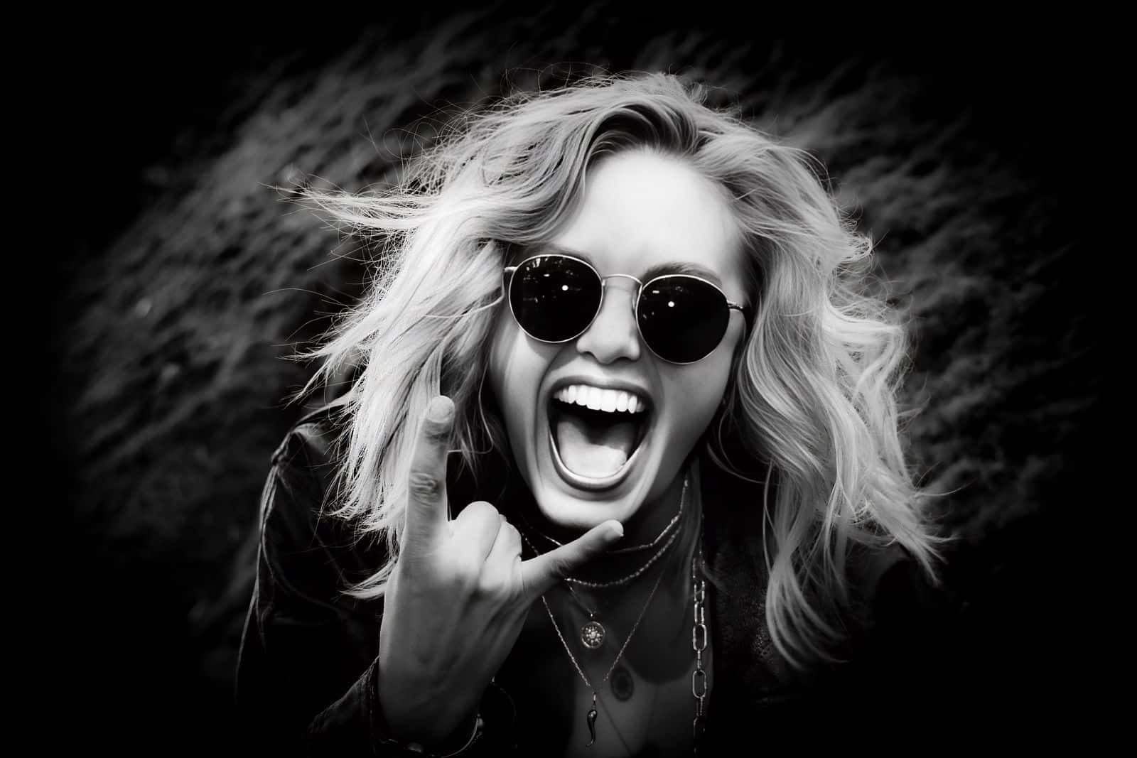 Black-and-white portrait of Mira laughing with her mouth wide open, wearing round dark sunglasses and layered necklaces, playfully making a shaka hand gesture against a dark natural background.