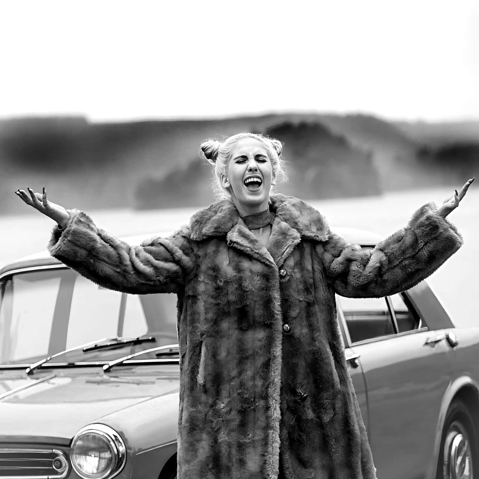 Peg Parnevik laughing with her arms outstretched in a fur coat beside a vintage car at an abandoned bathing spot south of Linköping, Sweden, in December.