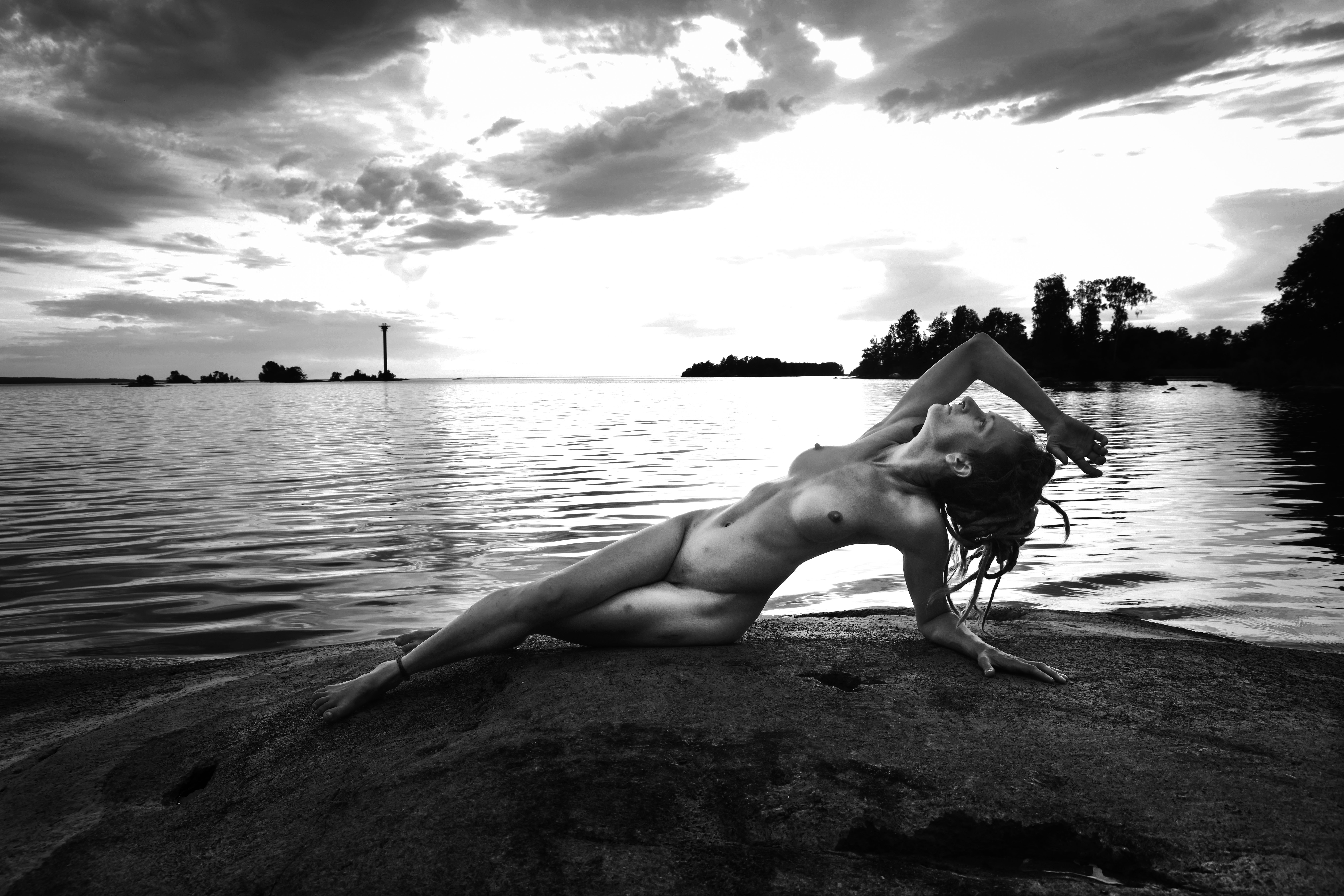A black and white photograph of a nude person posing gracefully on a large rock by the water. The person is reclining backward, supported by one arm, with the other arm elegantly arched over their head. In the background, calm water stretches toward the horizon under a dramatic, cloudy sky, with distant islands and a small lighthouse visible in the far distance.