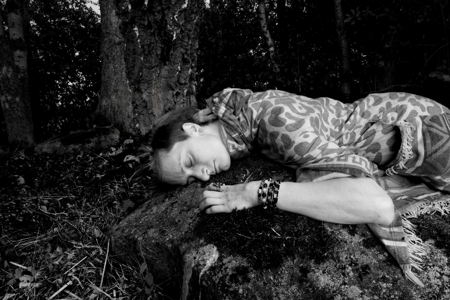 Black and white photograph of Smirja resting her head on a rock in the forest, wrapped in a patterned blanket.