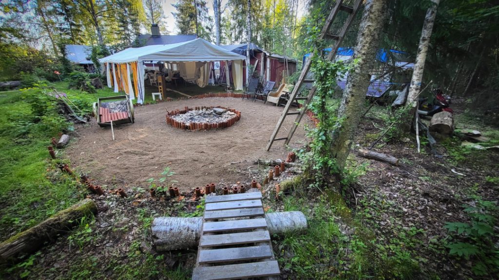 A group of naked people gathered around a fire in a forest clearing, sharing laughter, silence, and raw conversation — a moment of deep presence, honesty, and human connection at Rotum, a space where nothing is forced, and nothing needs to be explained.