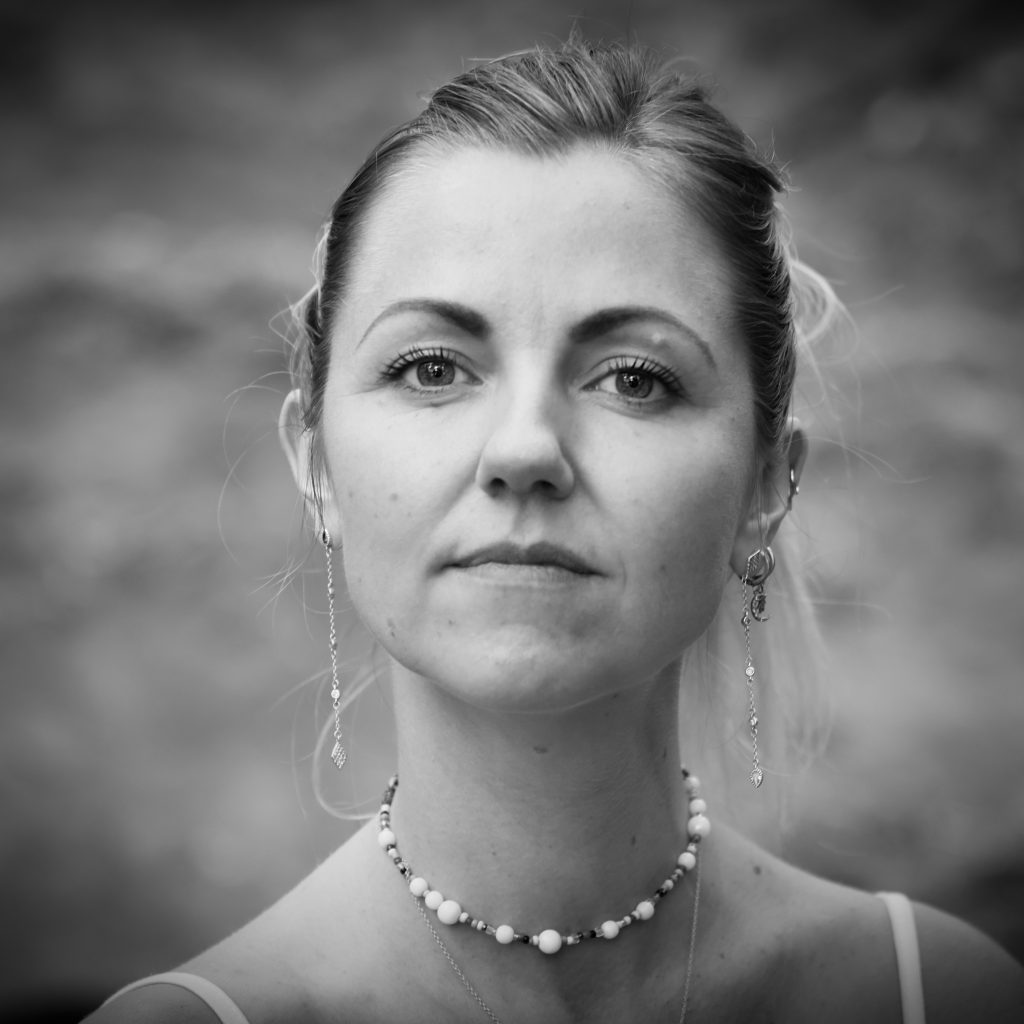 Porträtt i svartvitt av en kvinna med uppsatt hår, smycken och en pärlhalskedja. Hon ser rakt in i kameran med en lugn och närvarande blick
Black and white portrait of a woman with her hair tied back, wearing jewelry and a beaded necklace. She looks straight into the camera with a calm and present gaze.