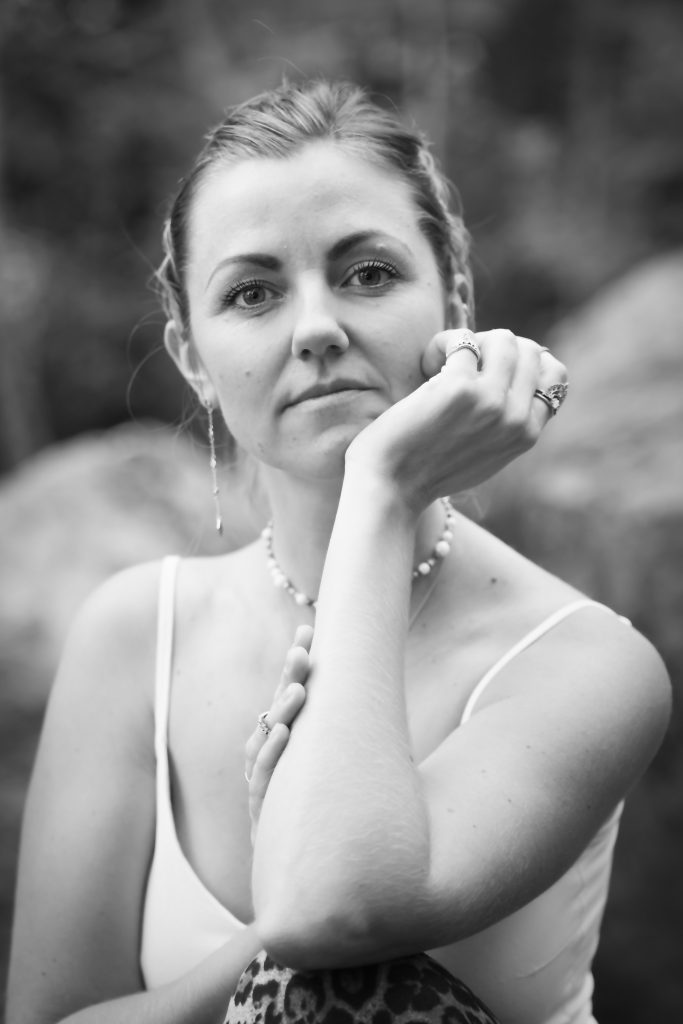 Black and white portrait of a woman with her hair tied back, wearing jewelry and a beaded necklace. She looks straight into the camera with a calm and present gaze.”