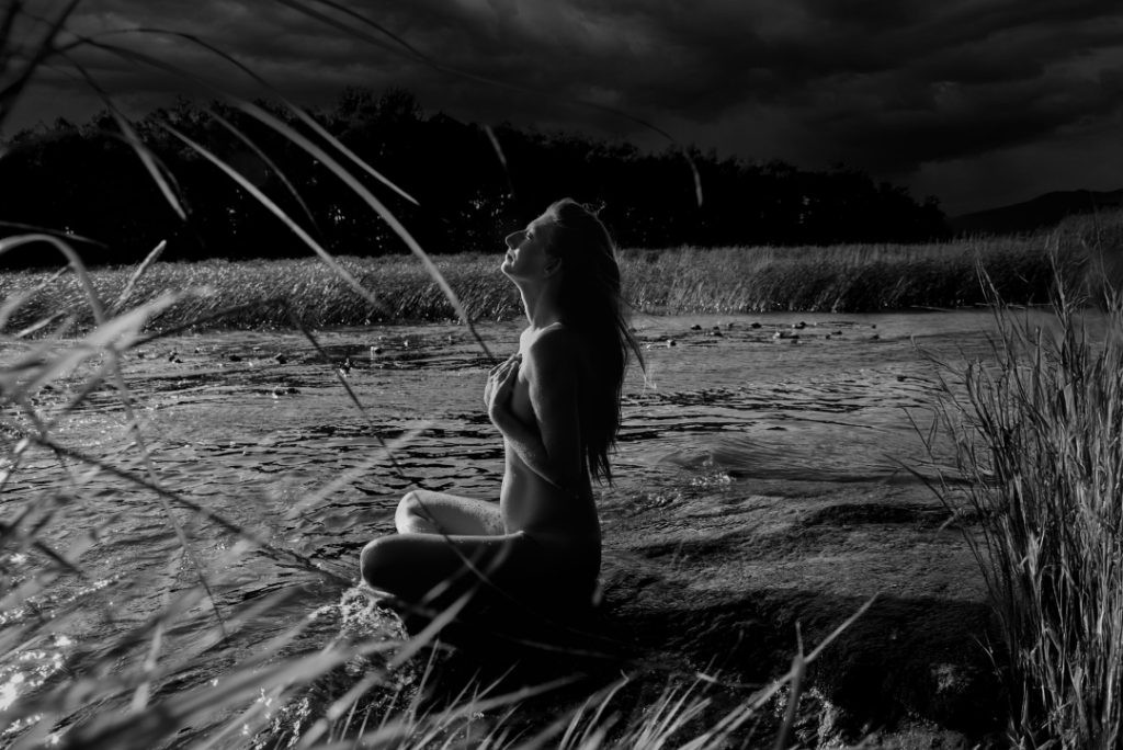 At the water’s edge, in the meeting between sky and lake, she seeks, nude, the light with closed eyes.