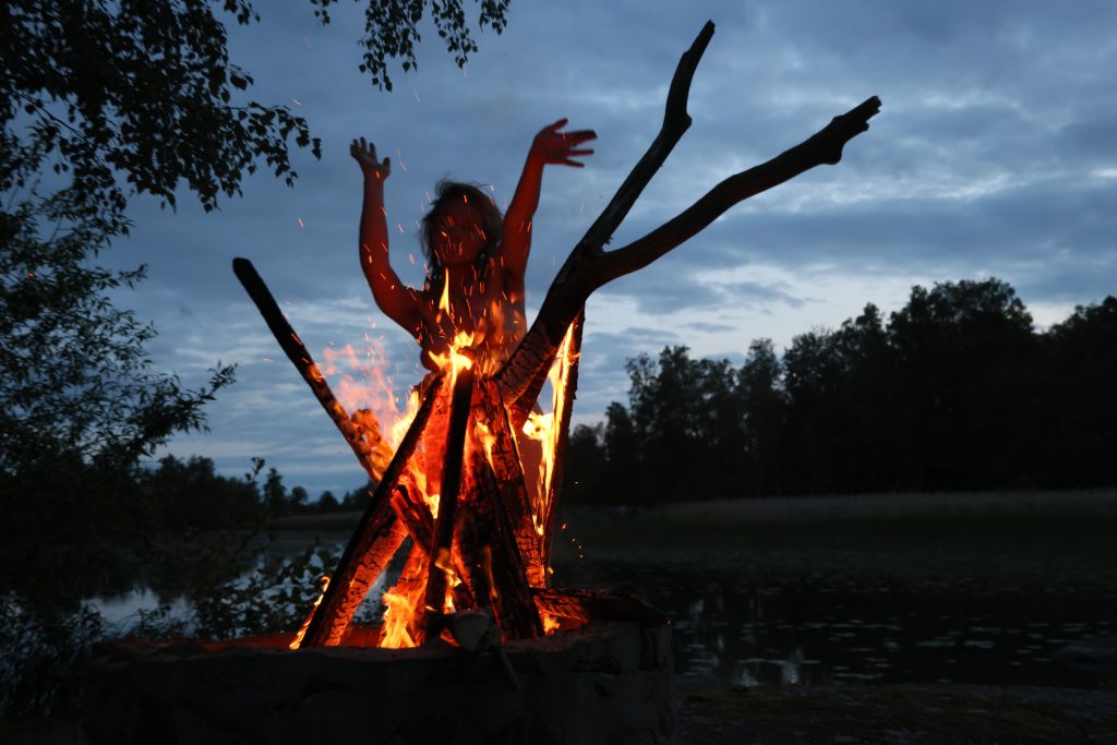 A woman stands behind a crackling fire at night, a lake faintly visible behind her. The glow of the flames illuminates her silhouette, conveying presence, strength, and quiet ritual in the dark landscape.
