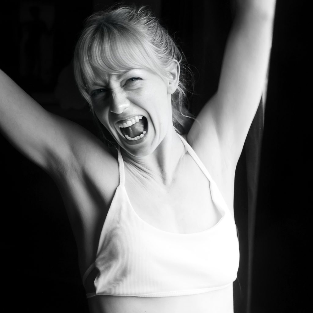 Porträtt av kvinna i vit topp som skrattar med armarna uppsträckta i mjukt studioljus.
